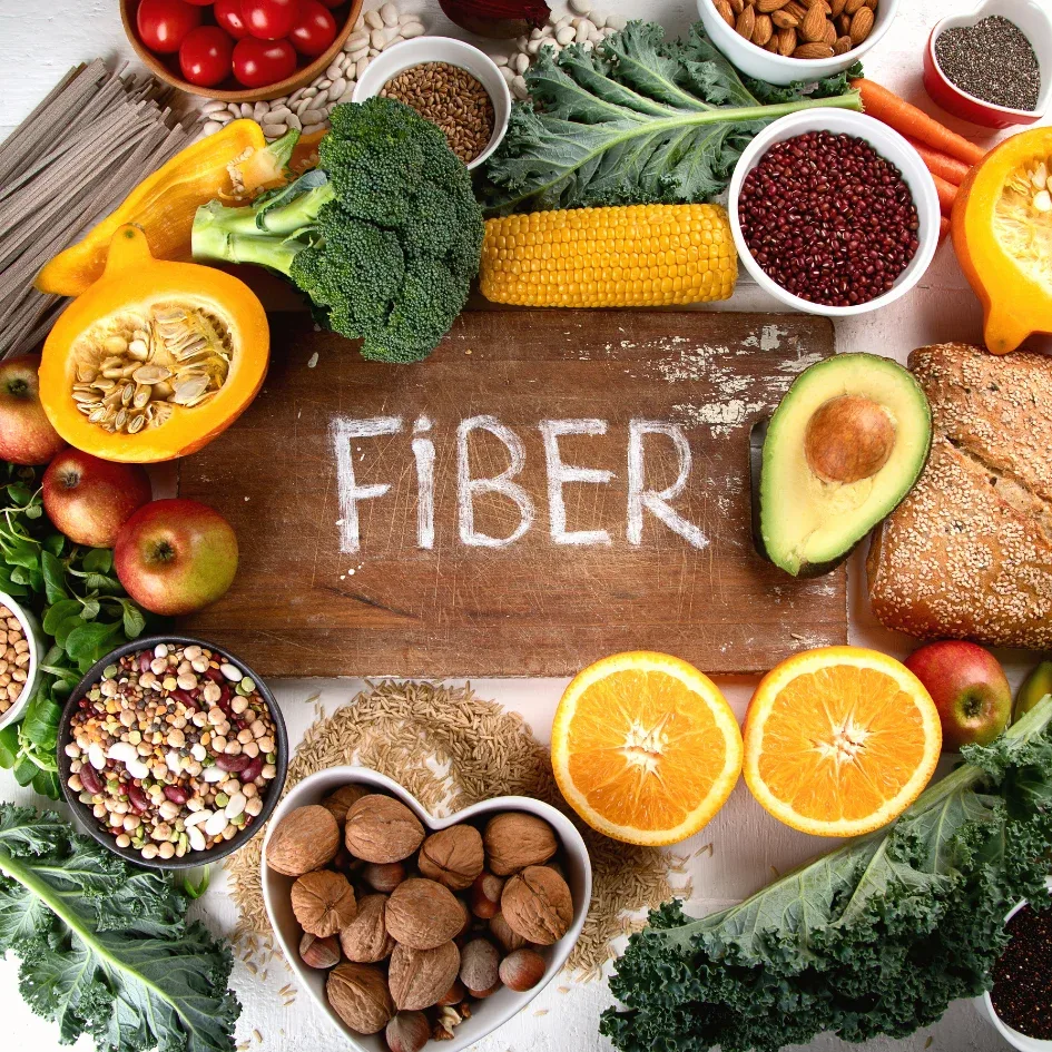 Imagen de una variedad de alimentos ricos en fibra, incluyendo vegetales, frutas, granos y nueces, dispuestos alrededor de una tabla con la palabra "Fiber" escrita en ella.