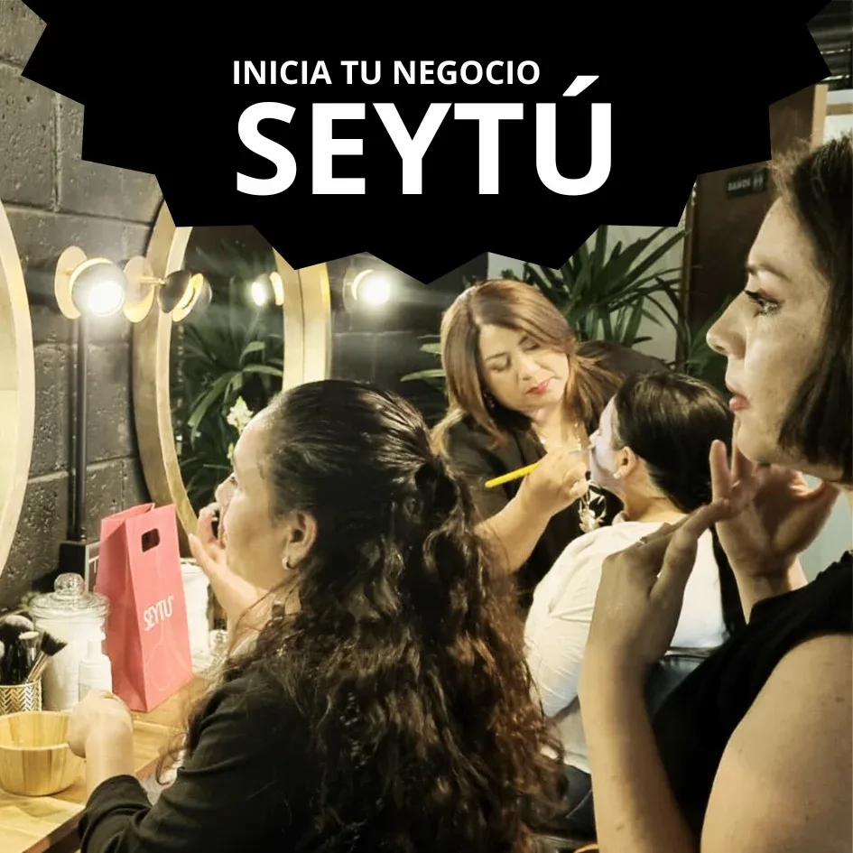 Mujeres aplicándose maquillaje en un salón de belleza con el texto 'Inicia tu negocio SeyTú' en la parte superior.