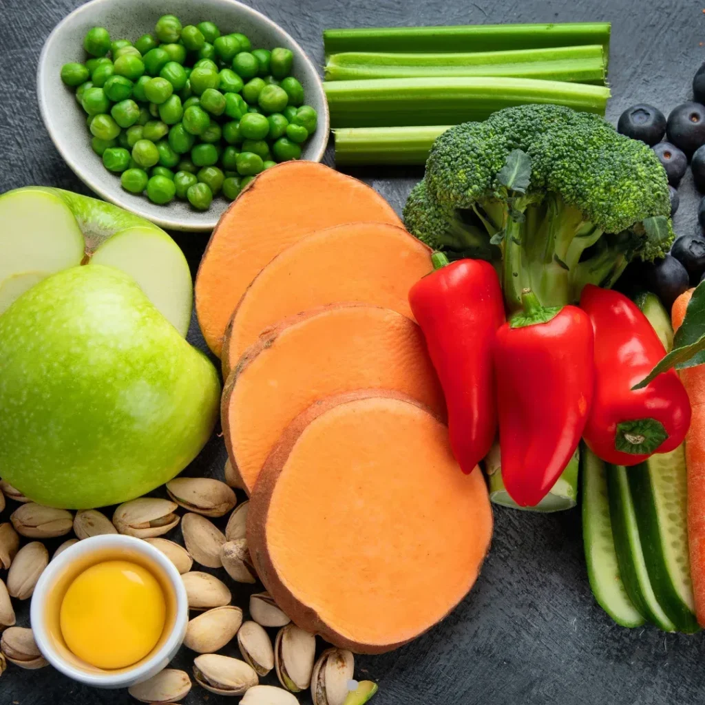 Variedad de alimentos saludables incluyendo camote, pimientos rojos, brócoli, apio, guisantes, manzana verde, pistachos y arándanos.