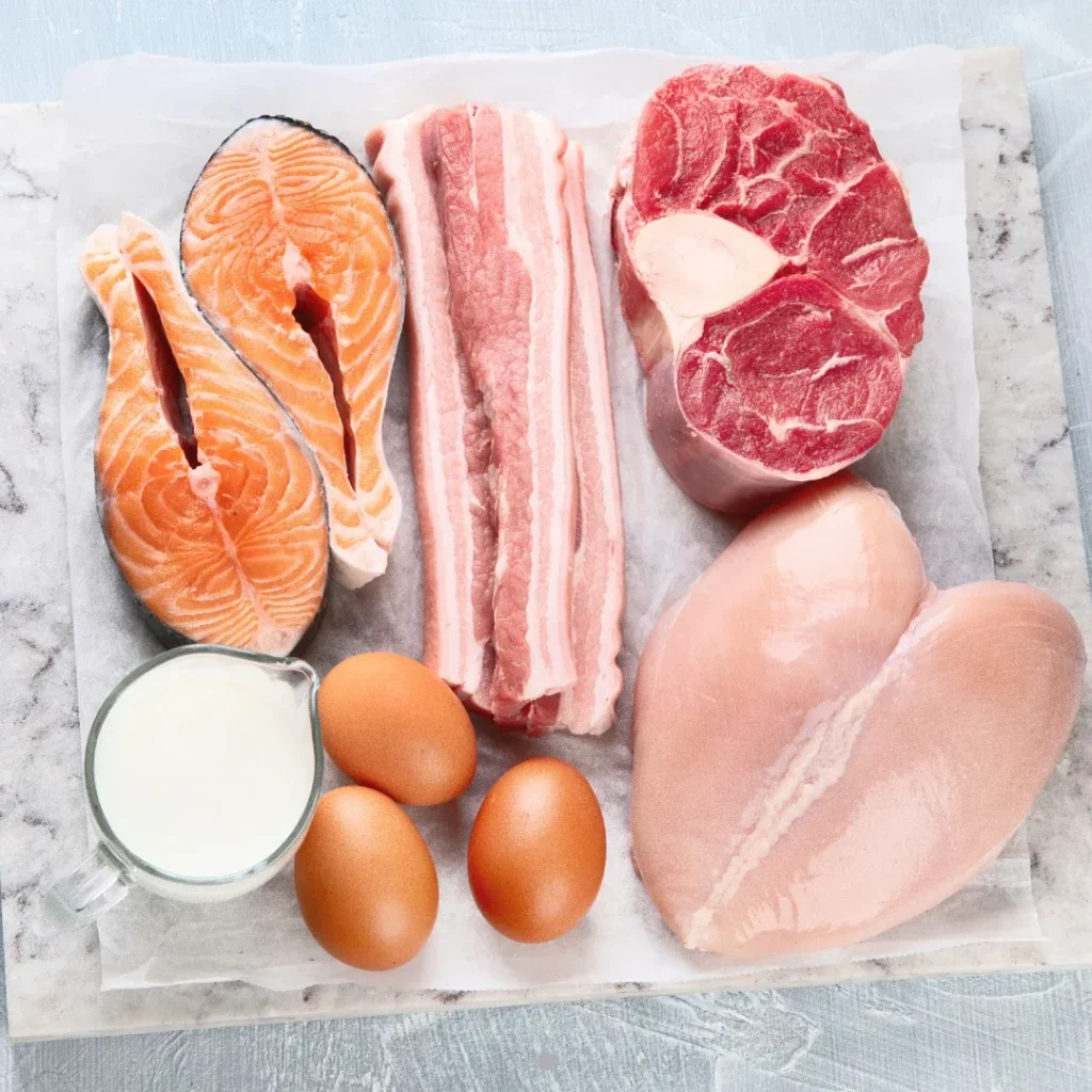 Diferentes tipos de proteínas animales, incluyendo salmón, tocino, carne de res, pechuga de pollo, huevos y leche, dispuestos sobre una superficie de mármol.