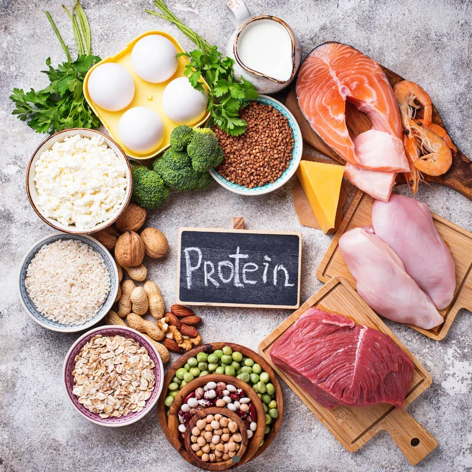 Diversas fuentes de proteínas, incluyendo carne, pescado, huevos, legumbres y nueces, dispuestas alrededor de una pizarra con la palabra "Protein".